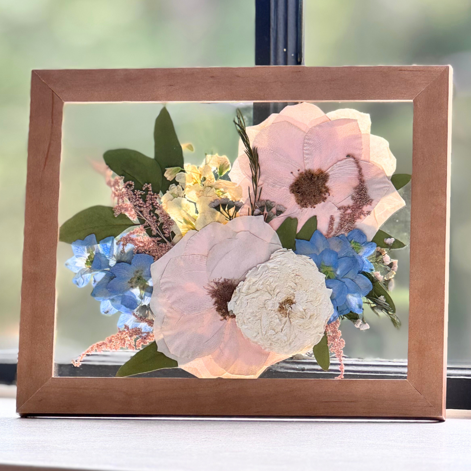 Freeze‑dried preserved wedding or funeral flowers artfully arranged in an 8 × 10 wooden frame. The composition includes soft pink and white blooms accented with blue florals and delicate foliage, offering a timeless keepsake of love and remembrance.