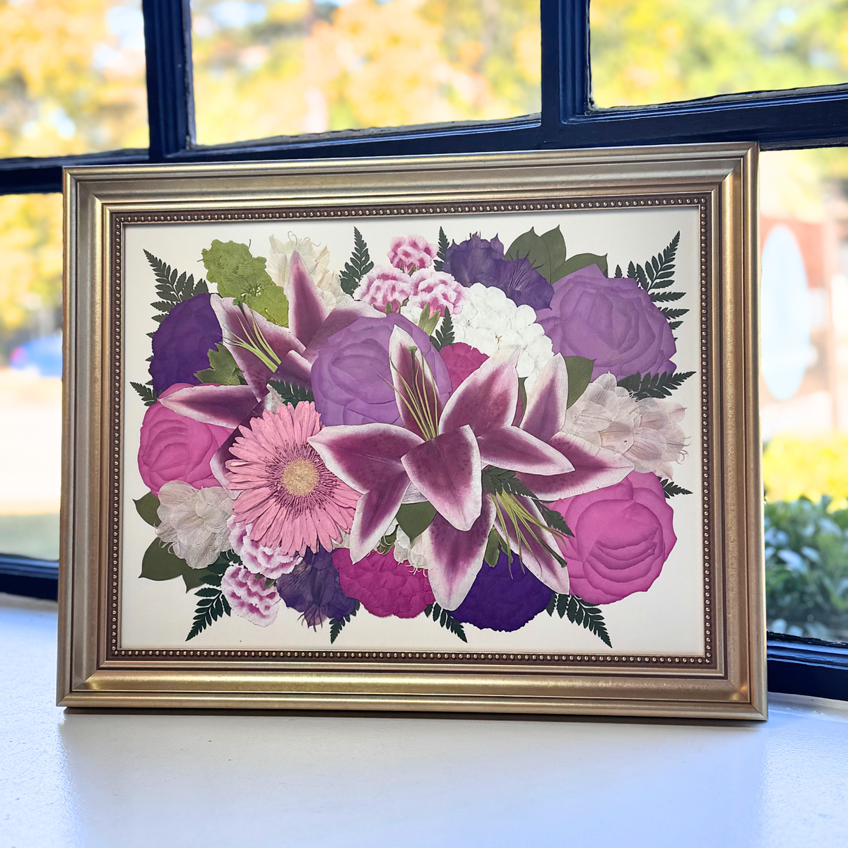 Framed 12x16 pressed flower art featuring deep purple lilies, pink roses, and assorted vibrant blooms, artistically displayed in a gold frame with window light.