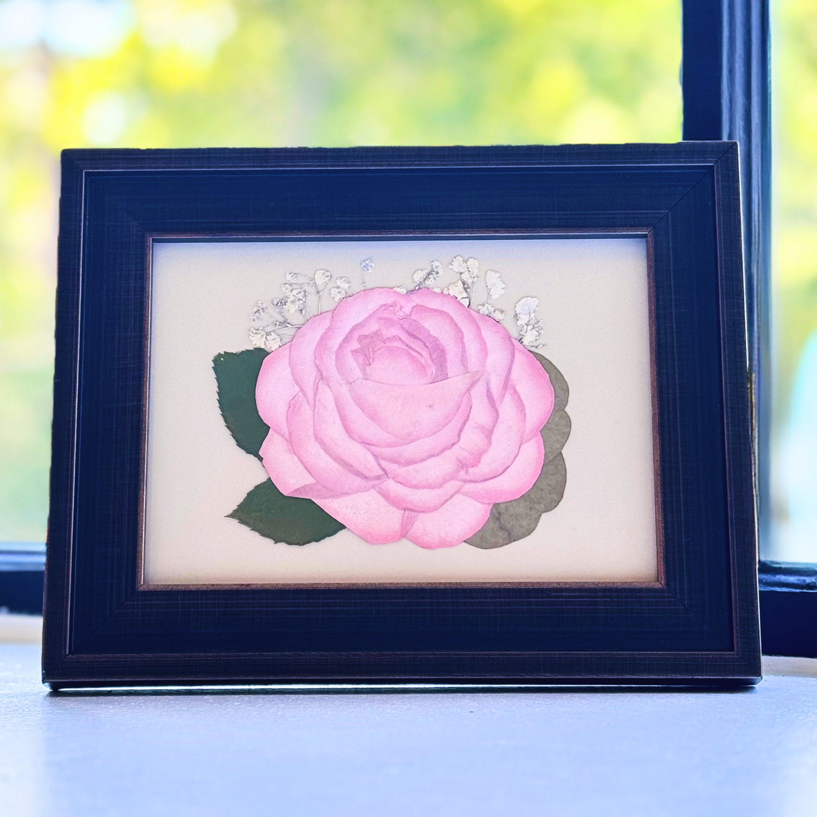 A single pink pressed rose displayed in a 5 × 7 dark wood frame, accompanied by sprigs of baby’s breath and soft green leaves. This gentle, romantic keepsake beautifully preserves a bloom from a wedding bouquet or memorial arrangement with timeless simplicity.