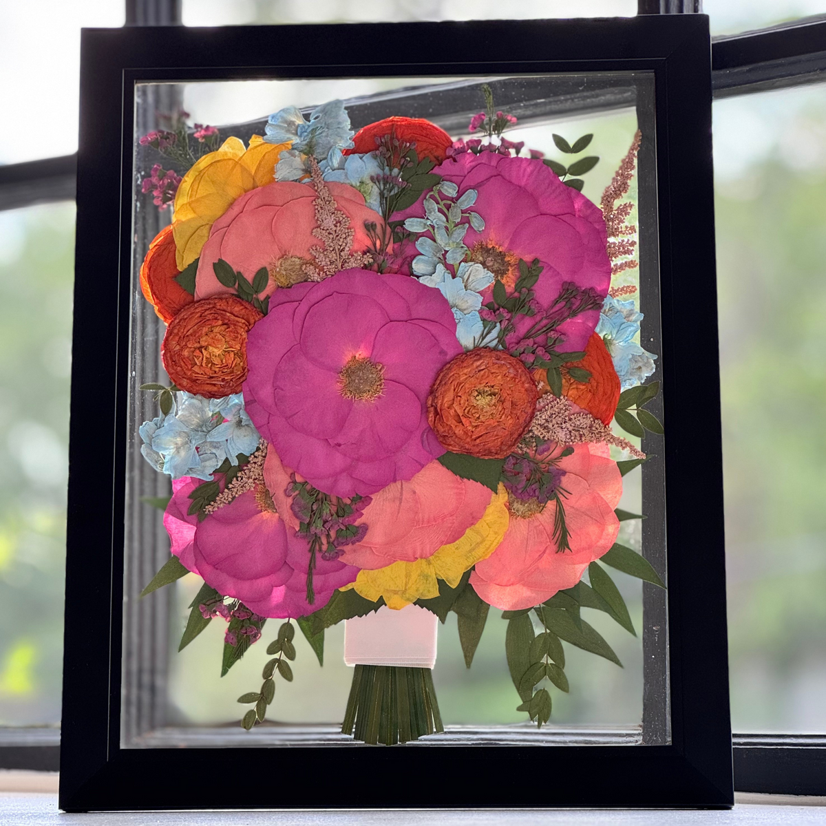 An 11 × 14 pressed flower frame capturing a vivid bouquet arrangement of hot pink, coral, orange, yellow, and blue florals with lush greenery, all tied at the base with a preserved ribbon stem. Displayed in a clear glass panel with a sleek black frame, this joyful keepsake preserves the energy and celebration of a wedding day.