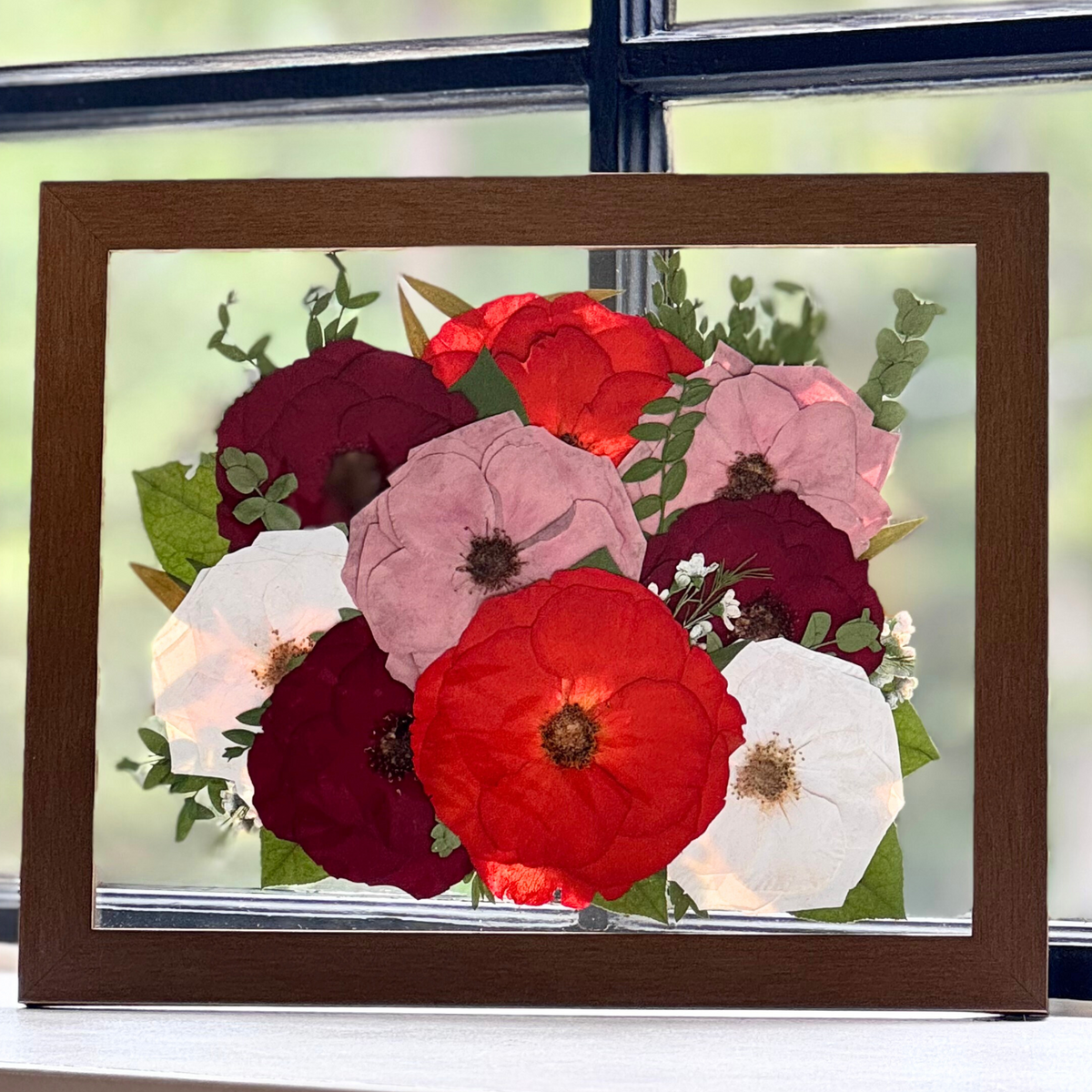 An 11 × 14 floating pressed flower arrangement featuring a lush mix of open roses in shades of deep red, blush, vibrant crimson, and ivory. Accented with greenery and baby&#39;s breath, this bouquet is encased in a warm wood frame—perfect for preserving the passion and elegance of a wedding or memorial floral tribute.