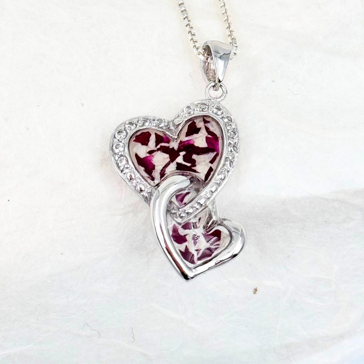 Sterling silver double heart pendant filled with preserved burgundy and white flower petals in resin, outlined with delicate crystal accents. A meaningful keepsake necklace created to celebrate the memory of a loved one.