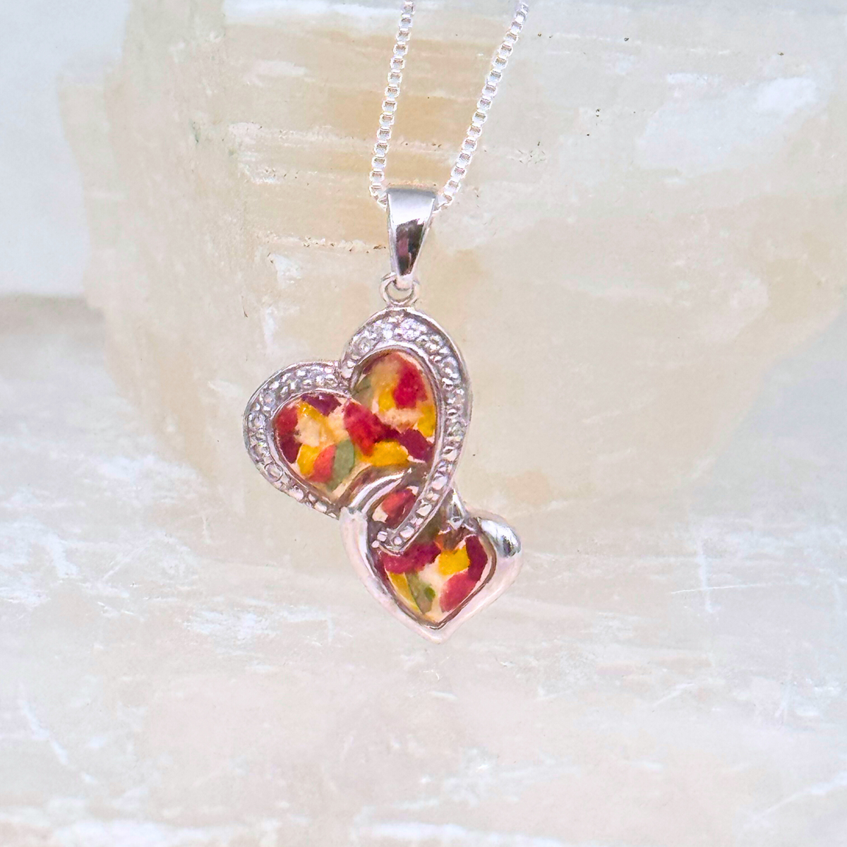 Sterling silver pendant featuring two interlocking hearts filled with preserved red and yellow flower petals in resin, framed with crystal accents. A beautiful memorial necklace to keep loved ones close.