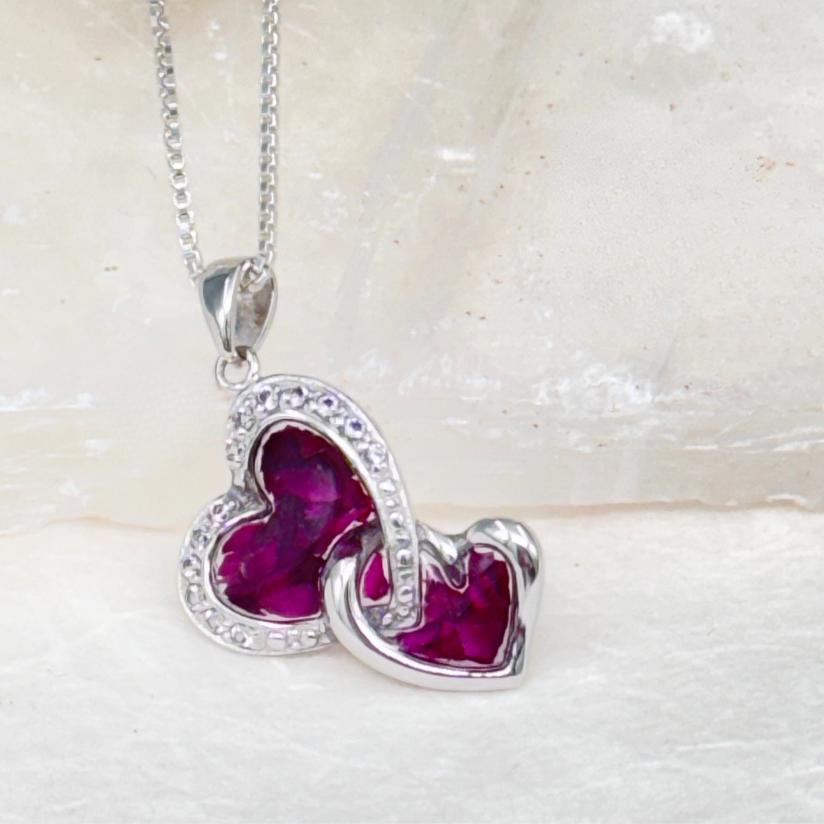 Sterling silver double heart necklace filled with vibrant deep pink preserved flower petals in resin, accented with shimmering crystal details. A touching piece of memorial jewelry for honoring a loved one.