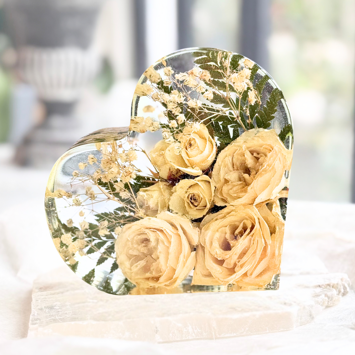 A handmade heart-shaped resin keepsake displaying preserved cream roses, baby's breath, and greenery, beautifully arranged in a clear resin block to commemorate a special occasion like a wedding or memorial.
