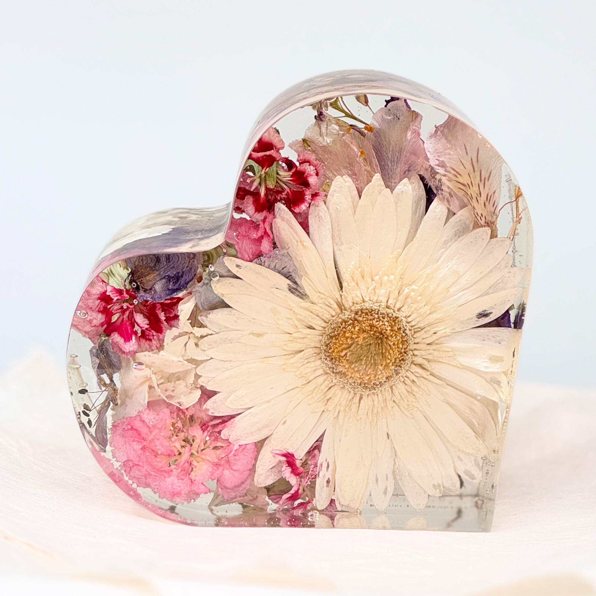 A heart-shaped resin keepsake featuring a white daisy surrounded by pink, purple, and red preserved flowers, handcrafted to display blooms from a special occasion like a wedding or memorial.