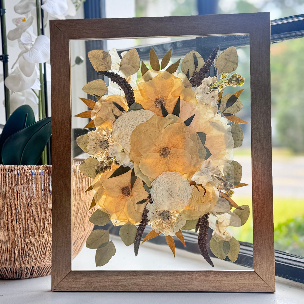 Floating 11x14 pressed flower art in wood frame with golden yellow, cream, and brown blooms on glass, displayed in natural window light