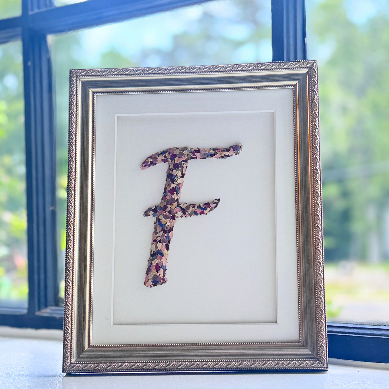 Custom floral monogram letter “F” made from preserved flower petals, framed in an 8x10 elegant gold frame, displayed by a window.