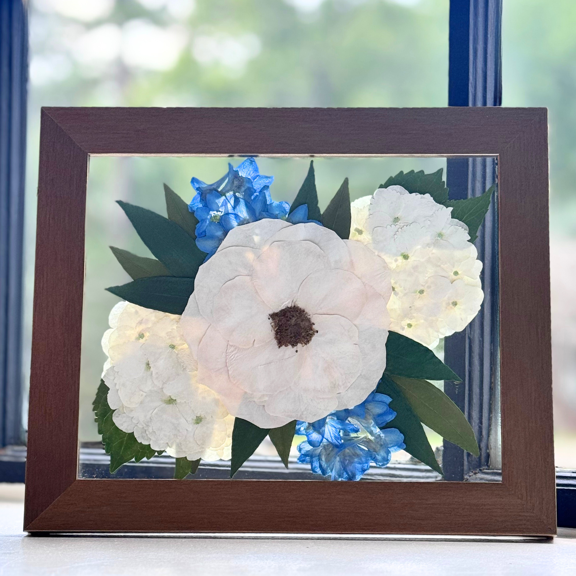 Pressed flower arrangement featuring a large white bloom surrounded by blue and white flowers, framed in a dark wood 8x10 shadow box. A handmade memorial keepsake capturing preserved wedding or funeral flowers.