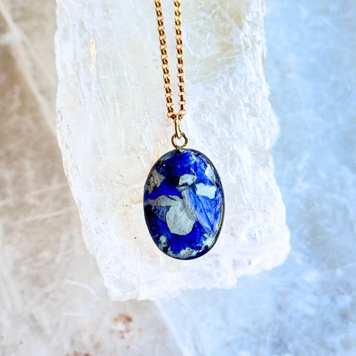 A gold-filled oval memorial pendant featuring a handmade inlay with preserved blue and white flowers, creating a timeless tribute.