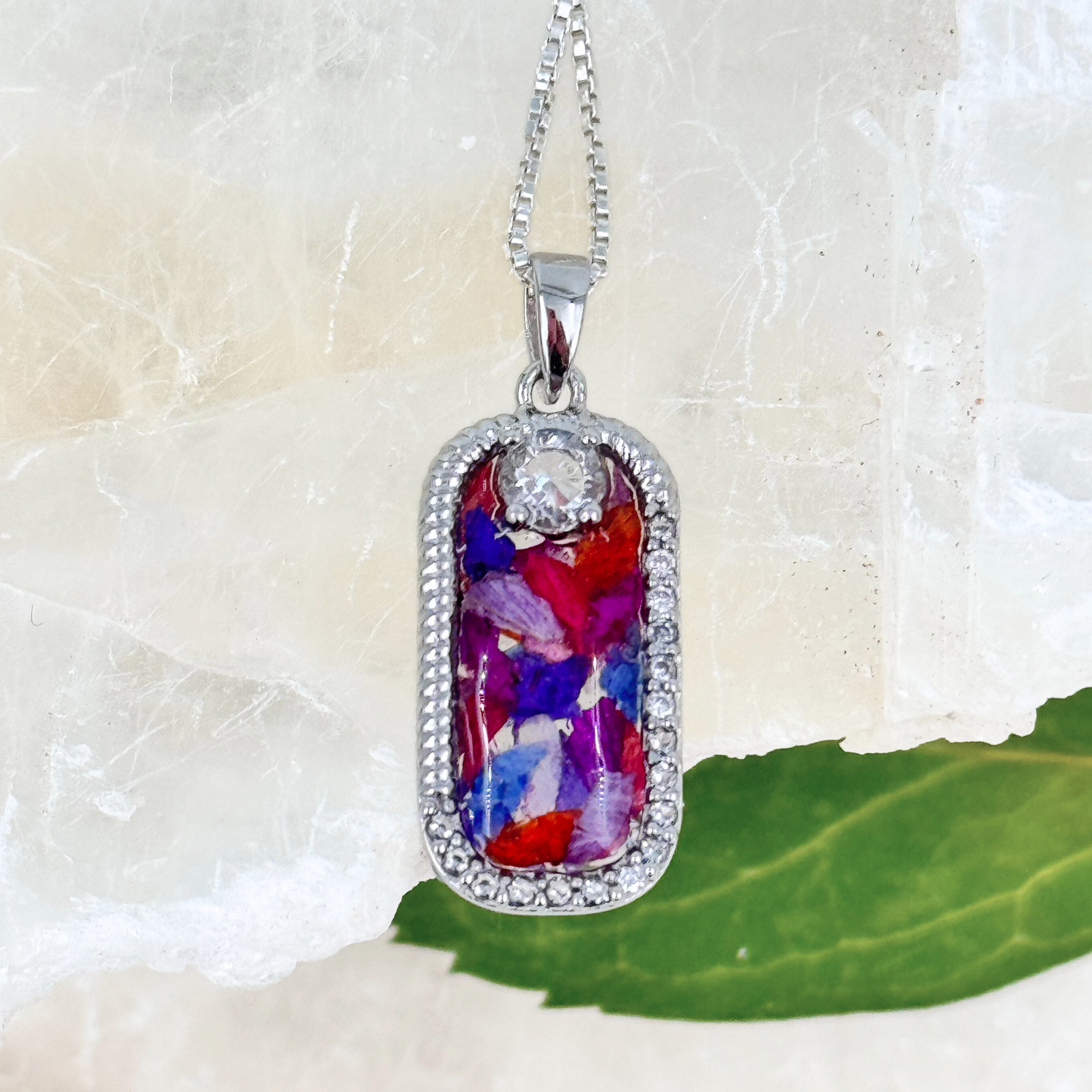 Sterling silver rectangular pendant featuring a mix of preserved red, purple, and blue flower petals set in resin, framed with delicate crystal accents and a single clear stone at the top. A memorial necklace created to honor loved ones.