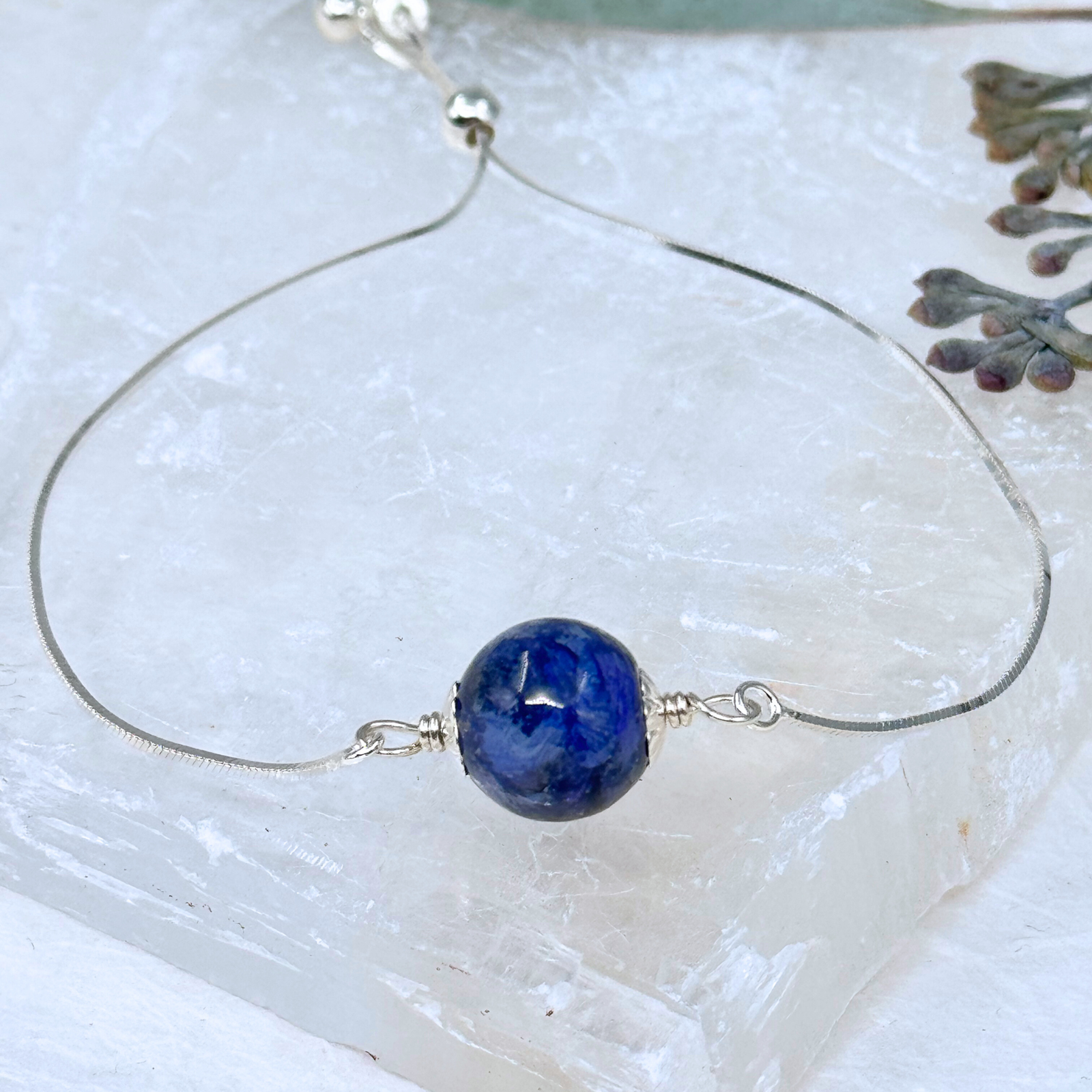 Close-up of a handcrafted memorial bracelet featuring a single bead made from preserved flowers, strung on a delicate sterling silver chain. The bead has a deep blue tone, reflecting the natural colors of the original petals.