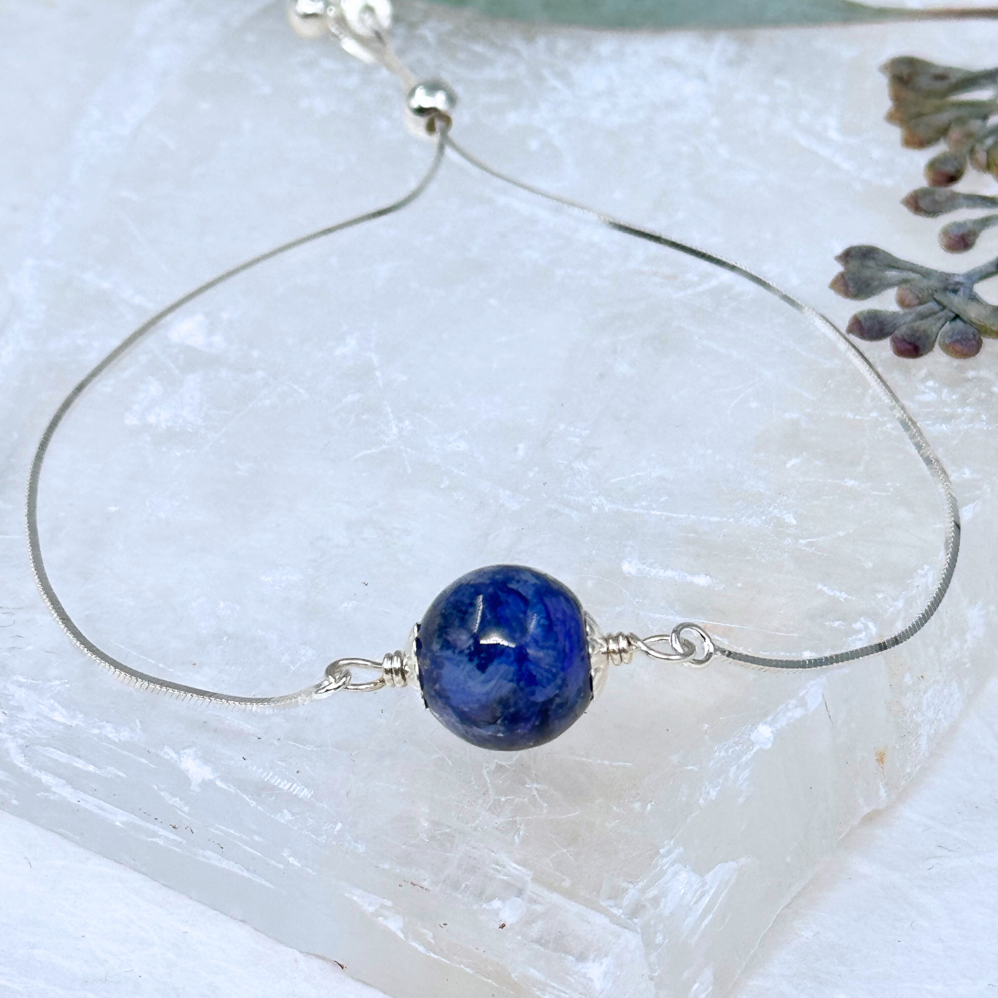 Close-up of a handcrafted memorial bracelet featuring a single bead made from preserved flowers, strung on a delicate sterling silver chain. The bead has a deep blue tone, reflecting the natural colors of the original petals.