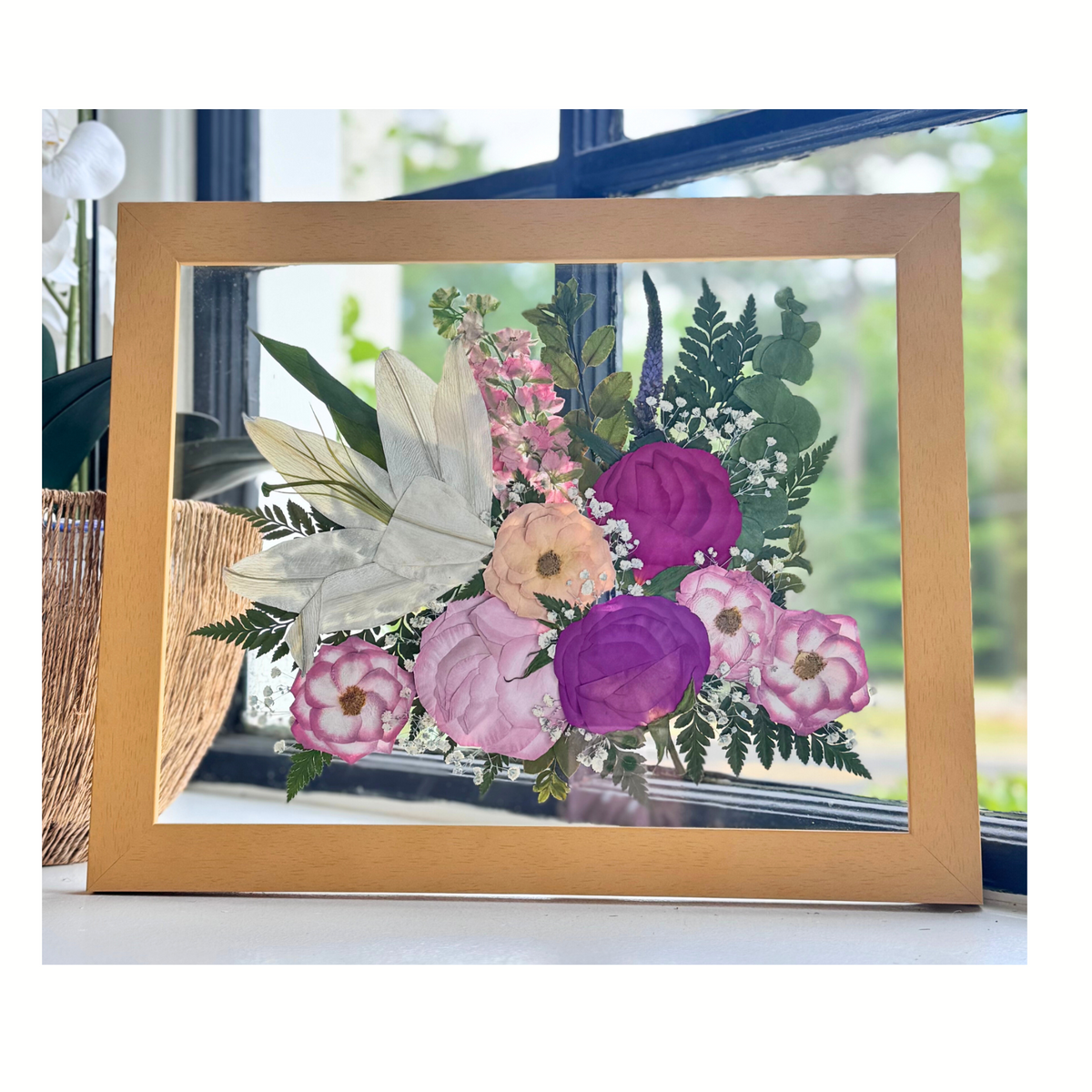 An 11 × 14 pressed flower arrangement featuring a white lily surrounded by blush, peach, and rich magenta blooms with baby&#39;s breath, ferns, and natural greenery. Framed in a light wood glass frame, this elegant piece beautifully preserves the soft romance of a wedding bouquet or a meaningful floral tribute.