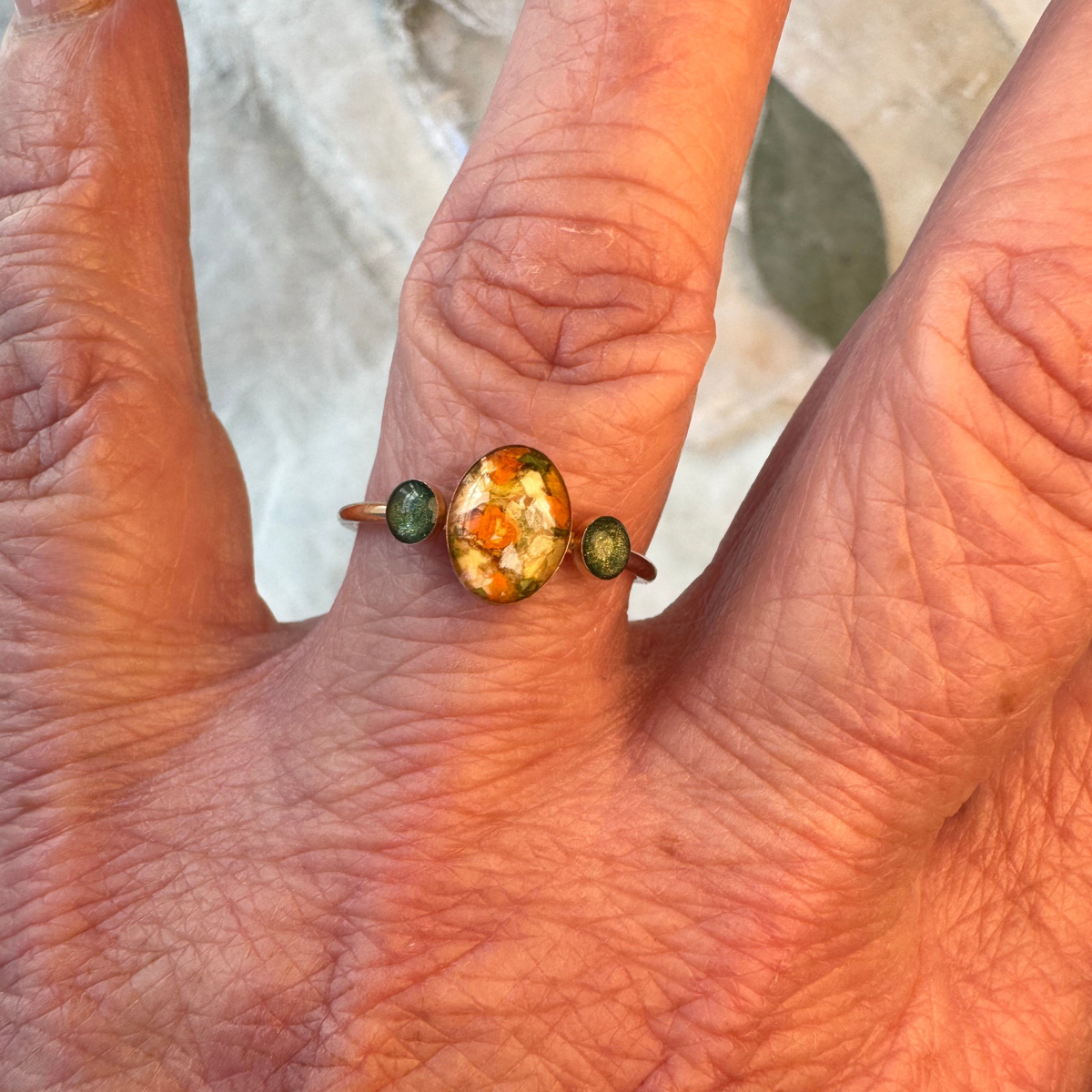 Meadowlight Keepsake Ring worn on a finger, featuring preserved wedding or funeral flowers in an 8x10mm oval center setting and two 5mm memorial settings for cremation ashes, handcrafted in gold-plated sterling silver.
