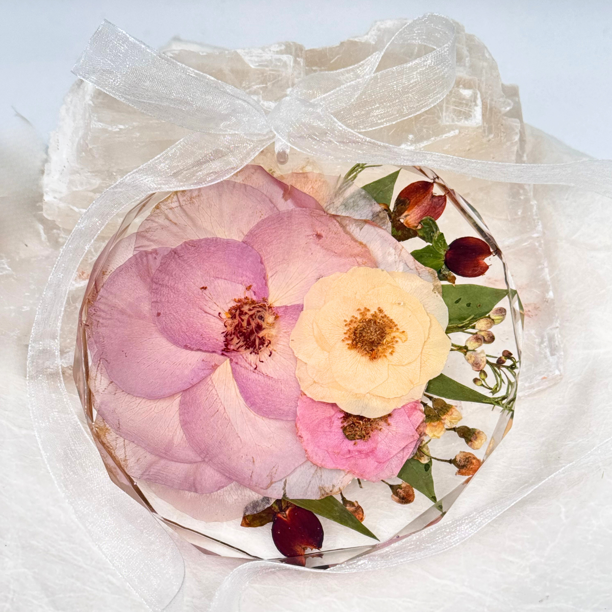 Round ornament made with preserved pressed flowers including pink, yellow, and burgundy blooms, tied with a sheer white ribbon for a delicate keepsake.
