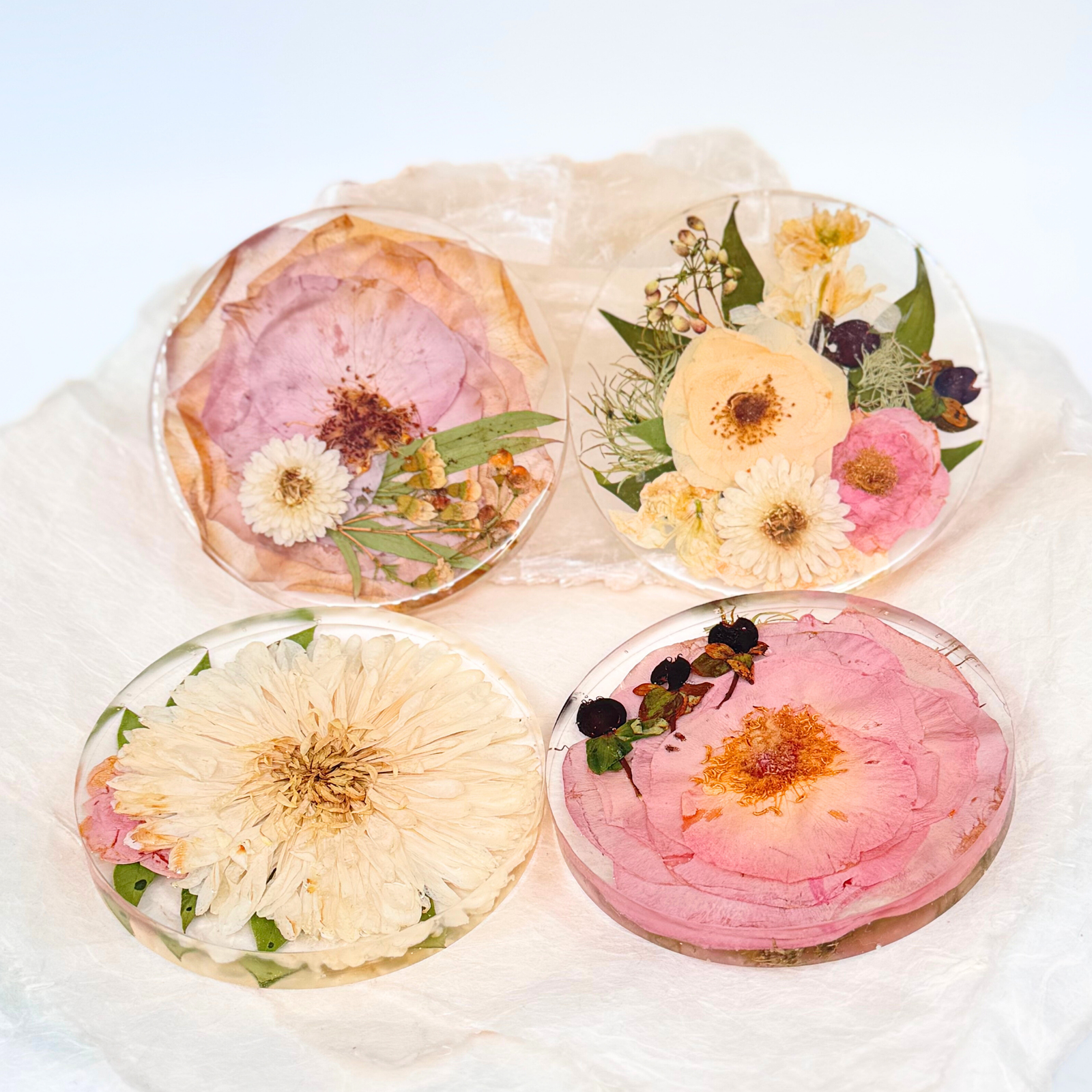 A set of four handmade resin coasters featuring preserved real flowers in various colors and arrangements, displayed on soft white fabric.