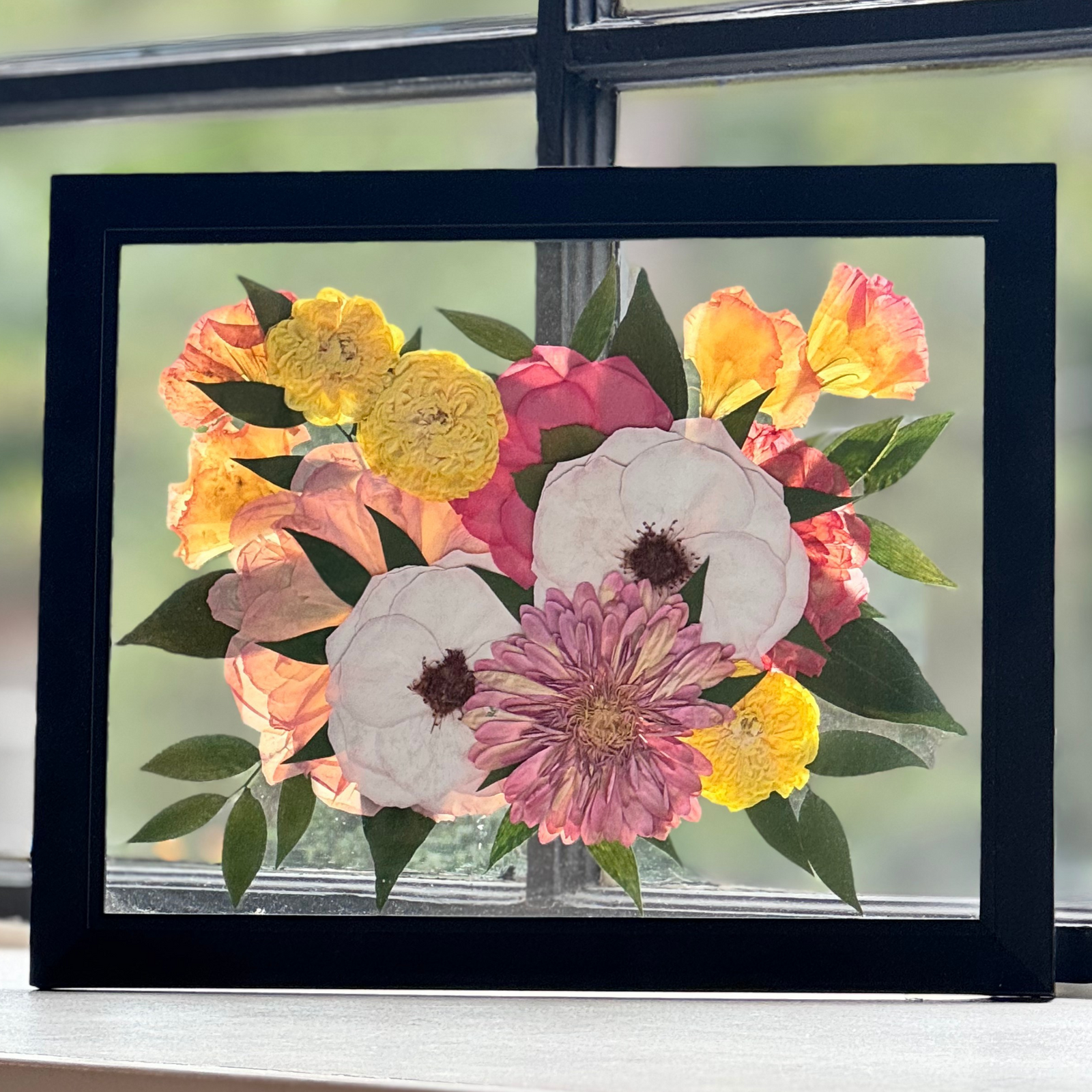 A 12 × 16 floating pressed flower frame featuring soft white open roses, yellow ranunculus, peach and coral blooms, and a pink daisy-like center flower. Surrounded by vibrant greenery and framed in sleek black, this radiant design evokes joy and celebration—ideal for preserving a wedding bouquet or bright memorial arrangement.