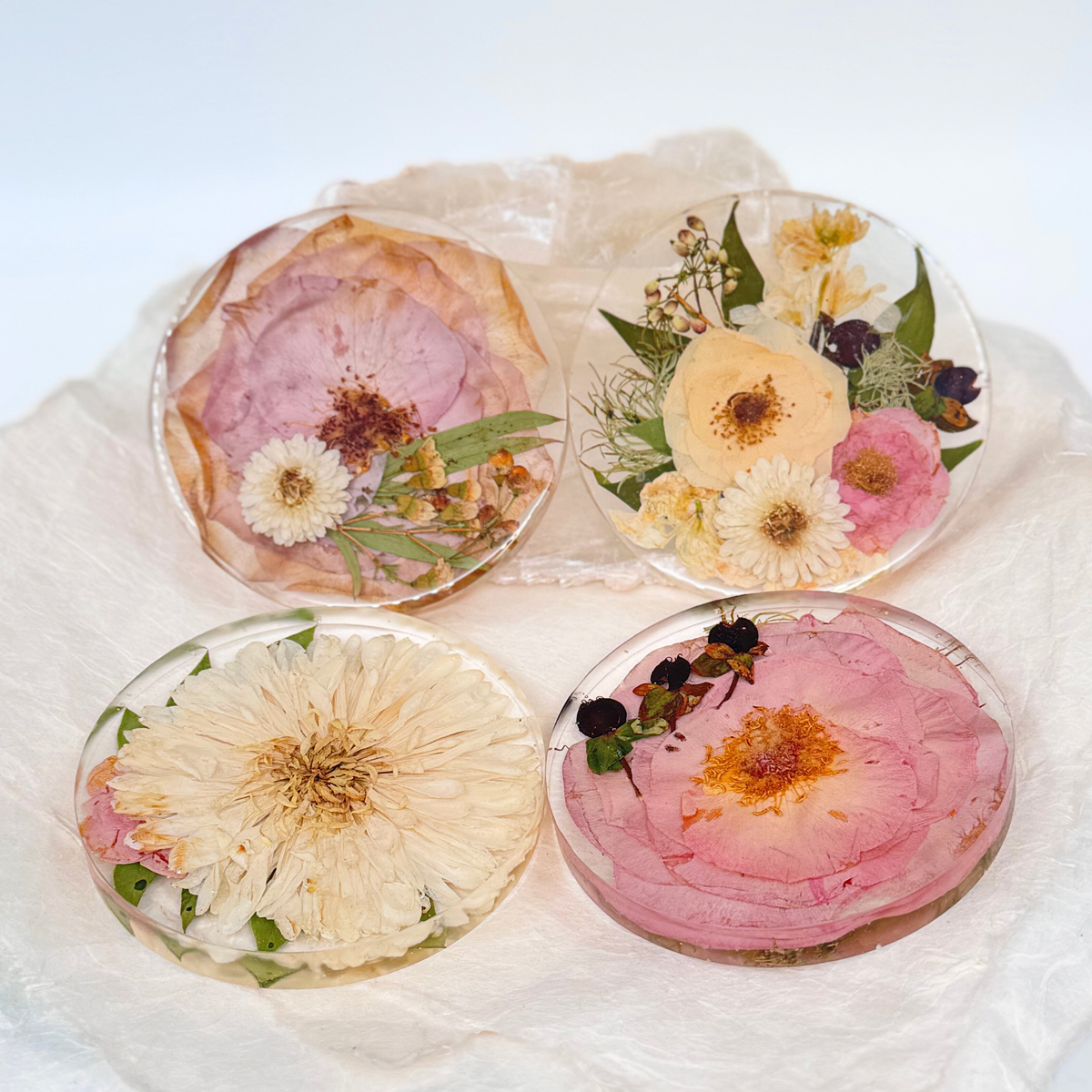 Set of four round resin ornaments featuring preserved flowers in soft tones of pink, peach, cream, and white, displayed on a white fabric background.