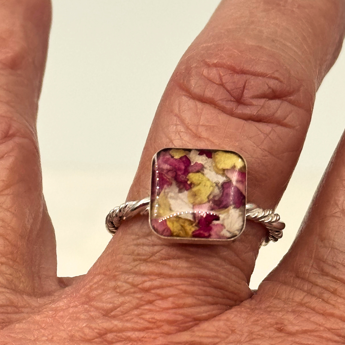 Sterling silver square memorial flower ring shown on hand, featuring preserved red, pink, and yellow funeral flowers in a resin setting with a twisted band.