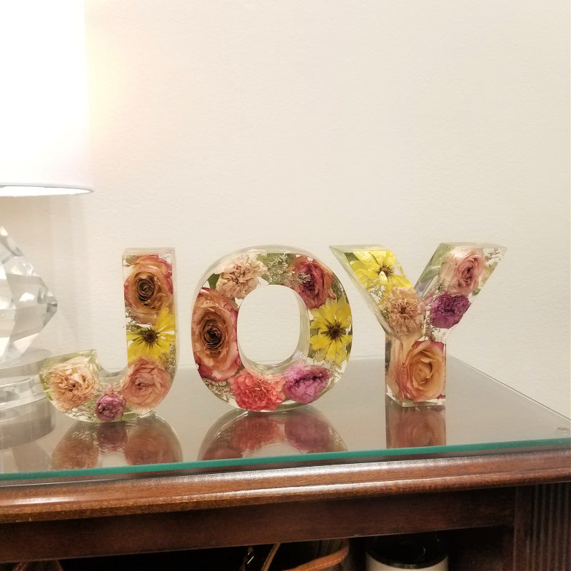 Set of resin letters spelling JOY, filled with preserved flowers in yellow, peach, pink, and purple tones from weddings or memorials, custom floral decor