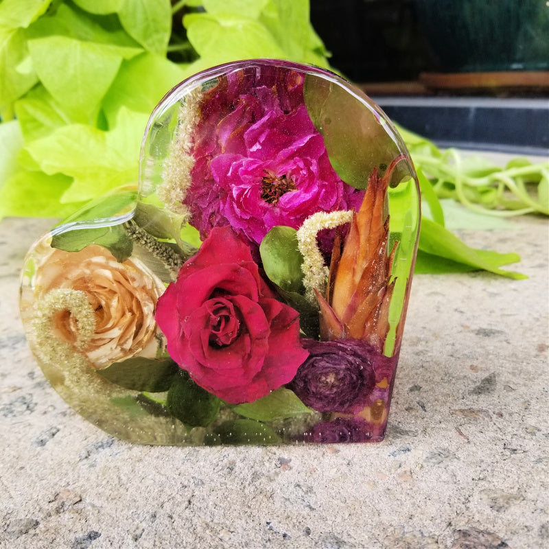 Heart-shaped resin keepsake filled with preserved bold flowers in shades of red, pink, peach, and orange, photographed on a stone surface with greenery in the background.