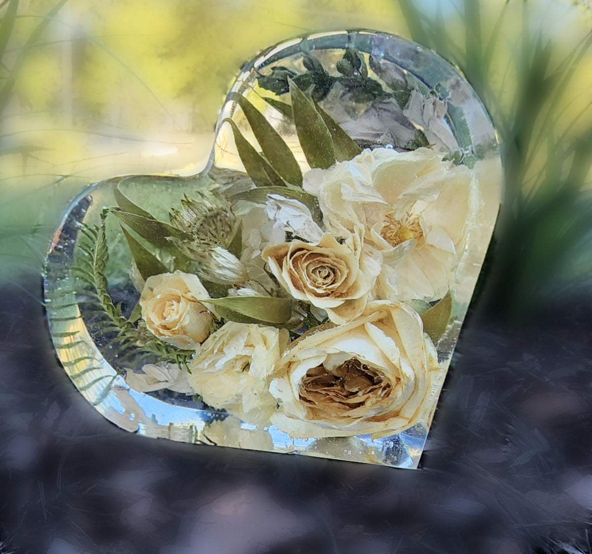 Heart-shaped resin keepsake featuring preserved pale yellow roses and greenery, photographed outdoors with soft natural light and a blurred garden background.