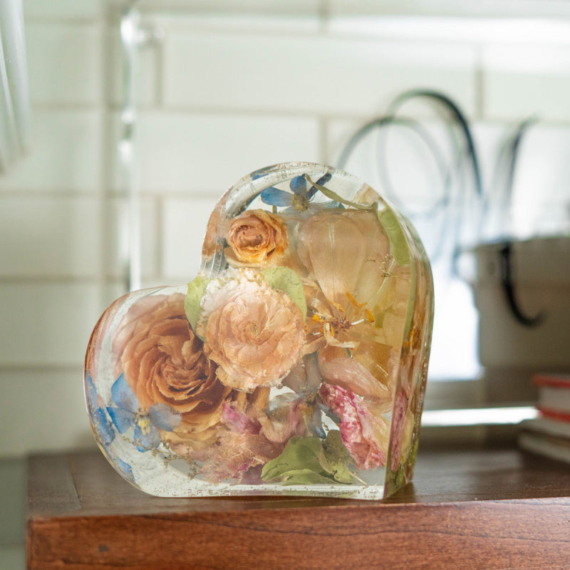 Heart-shaped resin keepsake with a mixed floral bouquet including roses, ranunculus, and greenery, displayed on a wooden surface with natural light.